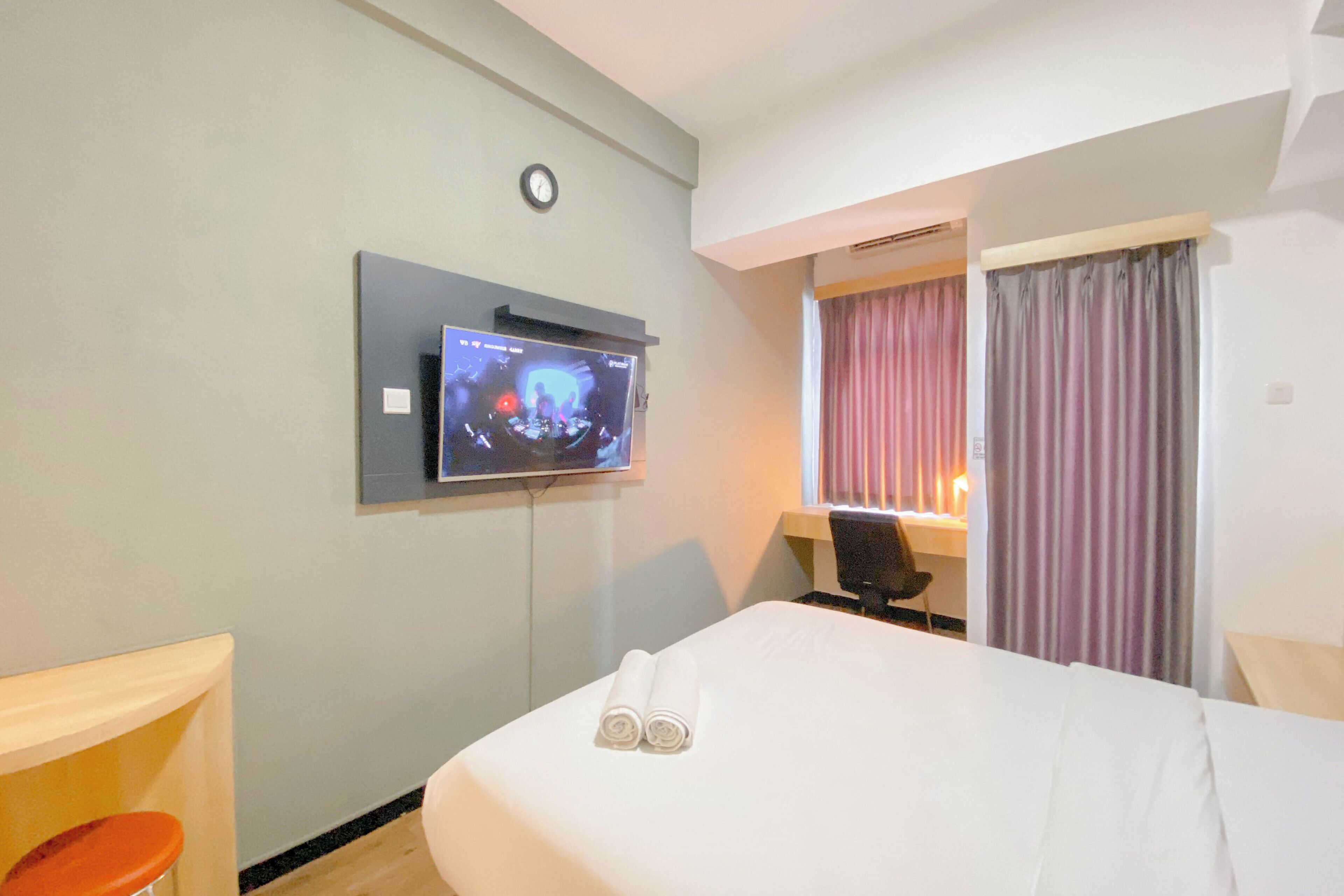 A hotel room with a bed, a TV mounted on the wall, a desk with a chair, and a window with curtains.