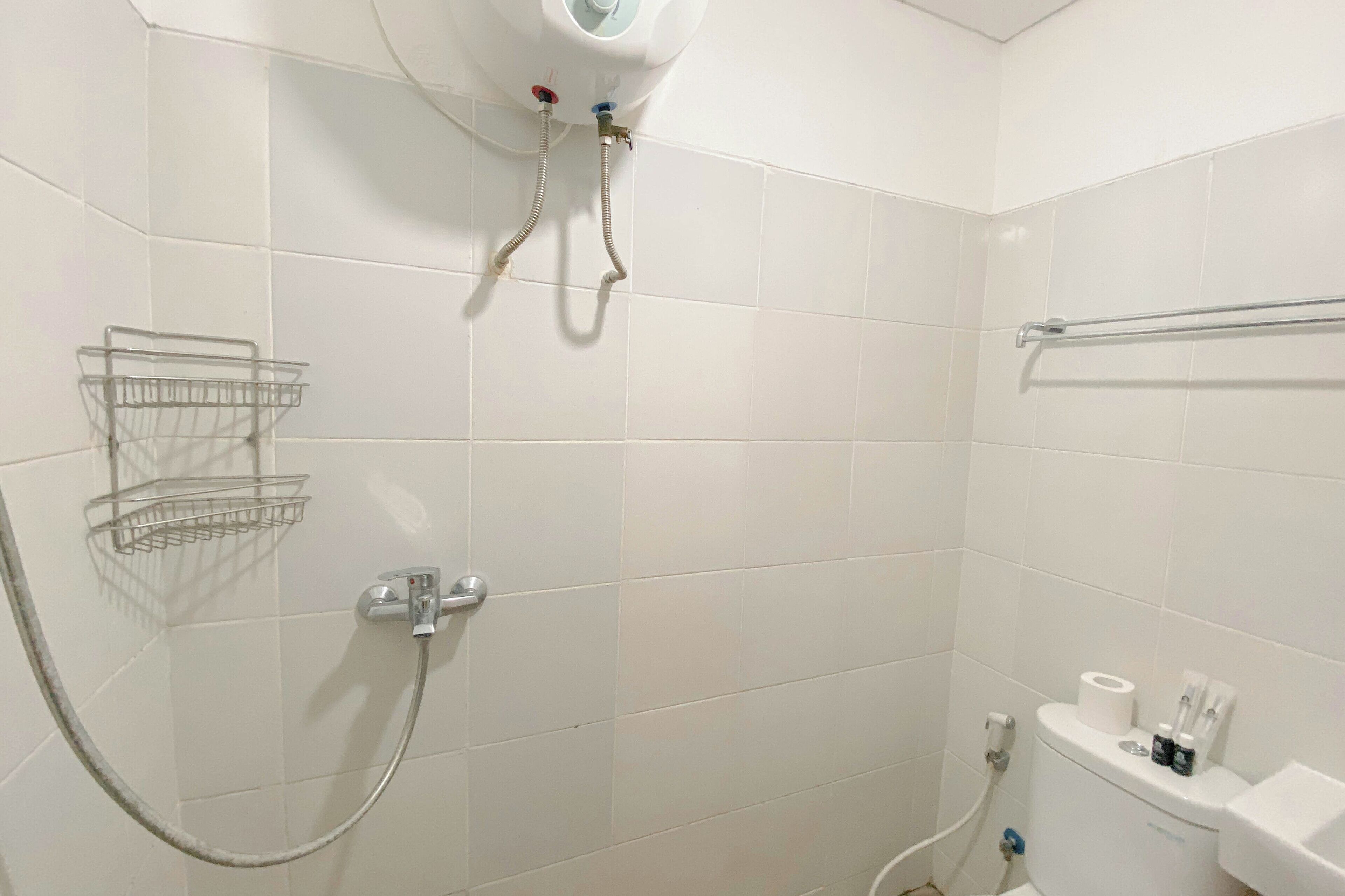 A bathroom with white tiles, a shower with a handheld showerhead, a towel rack, and a toilet with a paper towel dispenser.