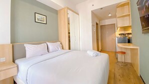 Une chambre d’hôtel avec un grand lit, une table de chevet en bois, un petit tabouret, un bureau avec une chaise et une kitchenette.
