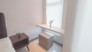 A small, neatly arranged room with a bed, a bedside table with a plant, a small stool, and a window with a flower vase.
