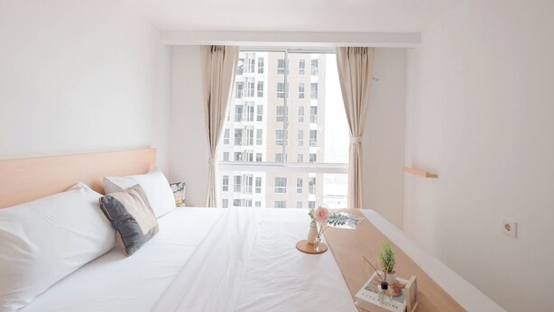 A hotel room with a large bed, a bedside table with a plant and a book, and a window with curtains. - Cozy And Spacious 2Br Apartment Tokyo Riverside Pik 2 (Teluknaga)