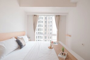 A hotel room with a large bed, a bedside table with a plant and a book, and a window with curtains. - Cozy And Spacious 2Br Apartment Tokyo Riverside Pik 2 (Teluknaga)