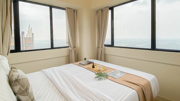 Een hotelkamer met een groot bed, twee ramen met gordijnen, uitzicht op een stadsbeeld en een kleine plant op het bed.