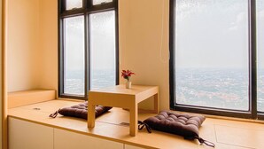 A corner room with a large window, a small table, two cushions, and a potted plant. - Japanese Style Studio At Pollux Chadstone Apartment (Cikarang)