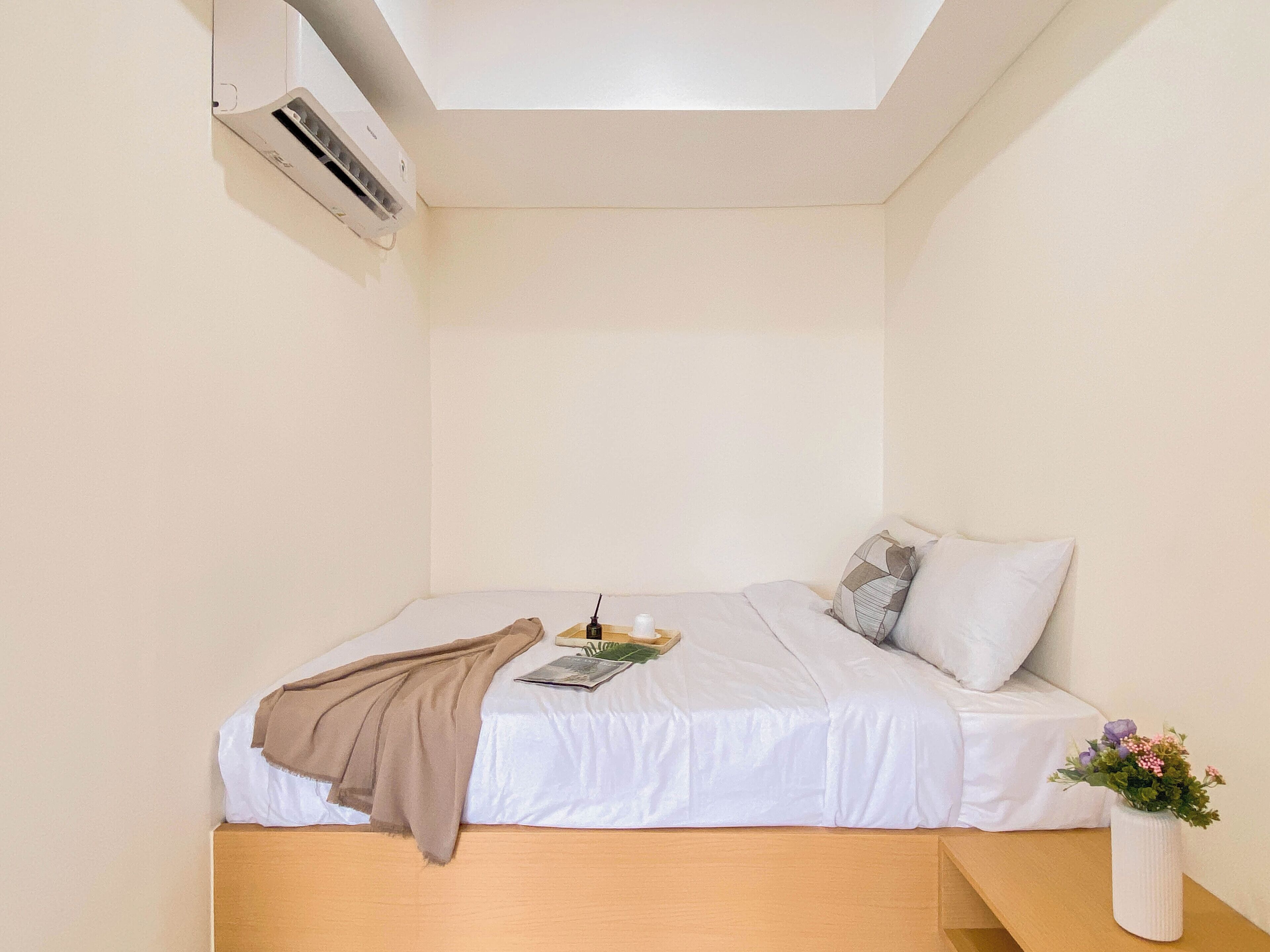 Ein kleines, ordentlich gemachtes Bett mit einer weißen Matratze und einer braunen Tagesdecke. Auf der rechten Seite des Bettes steht ein weißer Vasen mit Blumen.