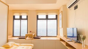 A modern living room with a sofa, a small table with a vase, a TV, and large windows offering a city view. - Japanese Style Studio At Pollux Chadstone Apartment (Cikarang)