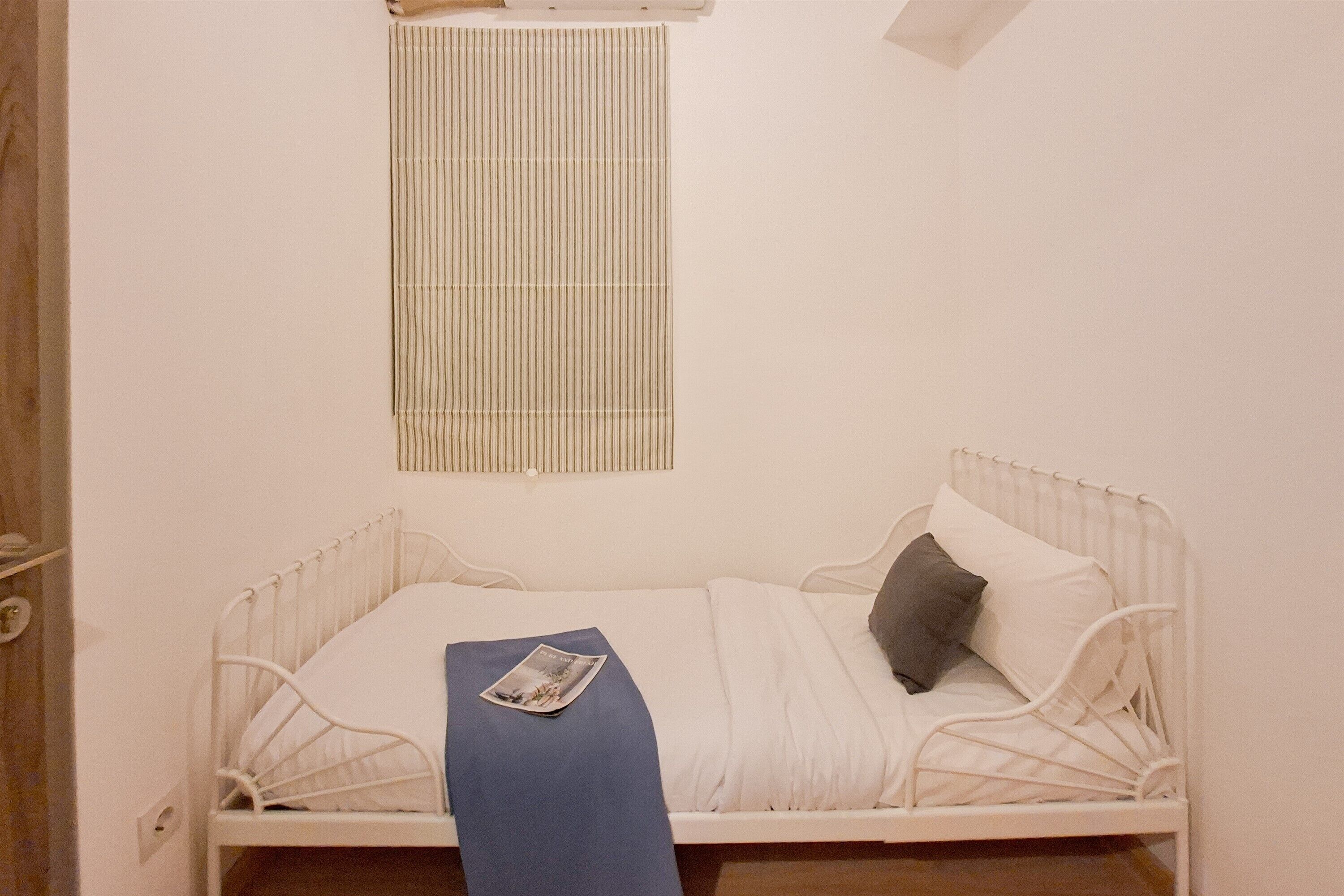 A single-bed room with a white metal frame, a window with vertical blinds, a bedside table with a magazine, and a door on the left.