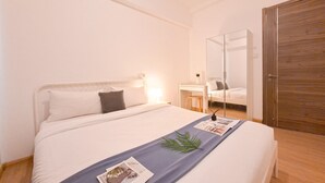 A neatly made bed with a blue bed runner, a bedside table with a lamp, and a large mirror on the wall. - Cozy And Pool View 2Br At Sky House Bsd Apartment (Pagedangan)