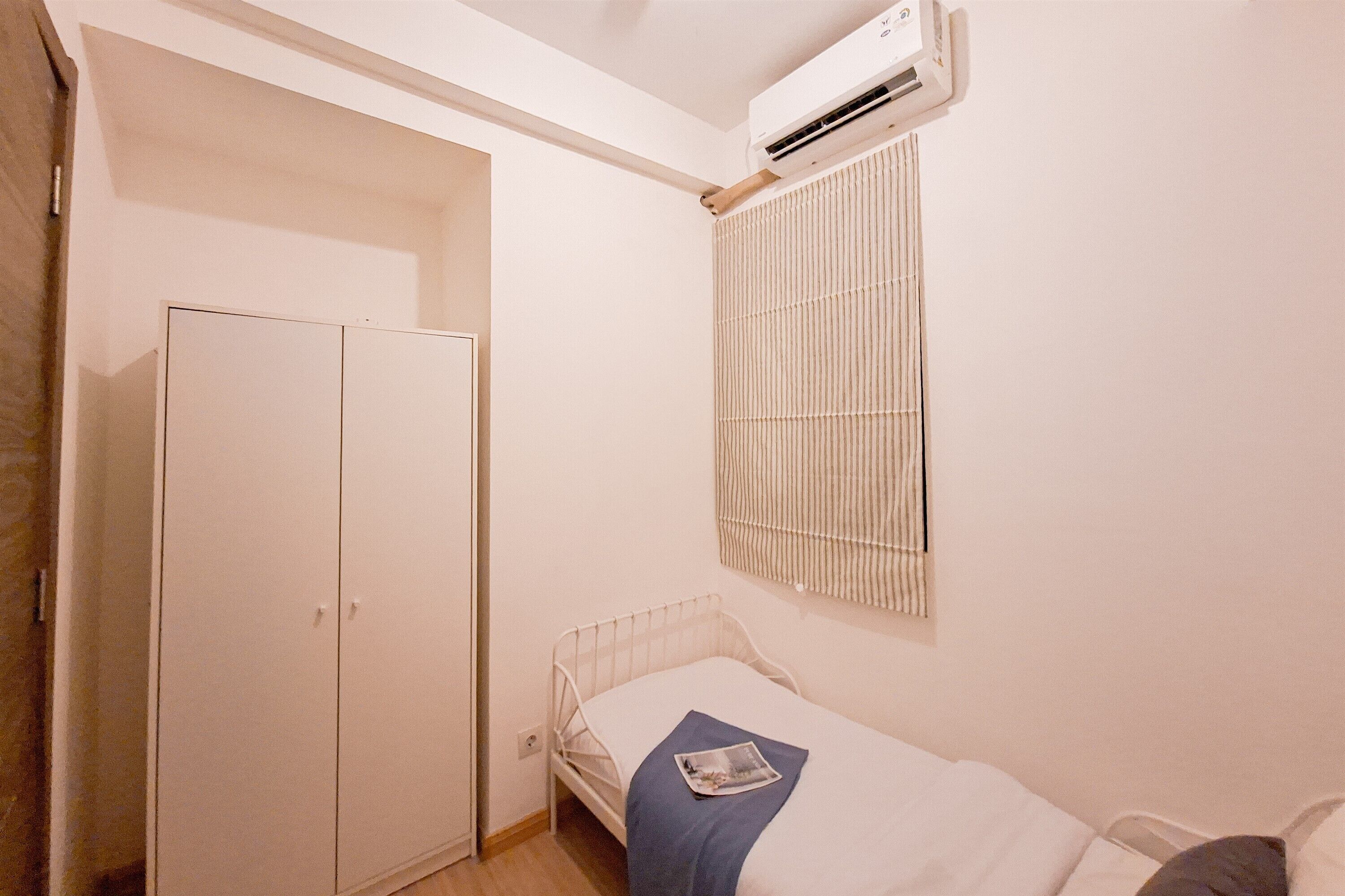 A small, single-bed room with a wardrobe, a window with blinds, and an air conditioning unit.