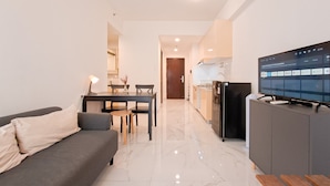 A modern living room with a sofa, a dining table with chairs, a kitchen area with a refrigerator, and a television on a cabinet. - Cozy And Pool View 2Br At Sky House Bsd Apartment (Pagedangan)