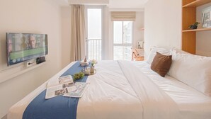 Habitación de hotel con cama, televisión montada en la pared, ventana con cortinas, escritorio con silla y estantería.