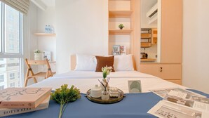 A hotel room with a neatly made bed, a small desk with a chair, a window with blinds, and a view of the city.