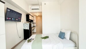 A compact hotel room with a bed, a small kitchenette, a TV mounted on the wall, and an air conditioning unit.