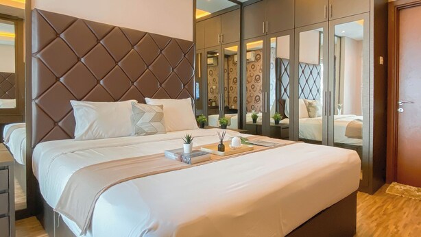 A modern bedroom with a large bed, a tufted headboard, a bedside table, and a wardrobe with mirrors. - Modern And Spacious 2Br Apartment Green Bay Condominium (Jakarta)