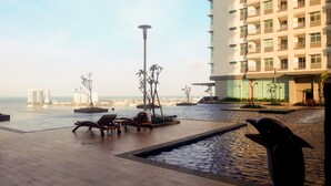 A rooftop pool area with lounge chairs, a dolphin sculpture, and a view of the sea and buildings. - Modern And Spacious 2Br Apartment Green Bay Condominium (Jakarta)