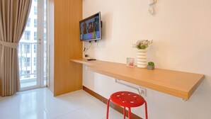Une chambre moderne avec un bureau en bois, un téléviseur fixé au mur, une petite plante en pot, un tabouret rouge et une fenêtre avec des rideaux.
