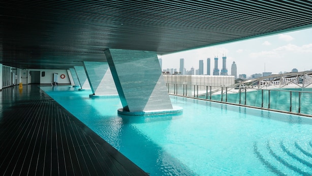 Une piscine intérieure moderne avec vue sur les toits de la ville, dotée d’une architecture élégante, d’une terrasse en bois et d’une rambarde en métal.