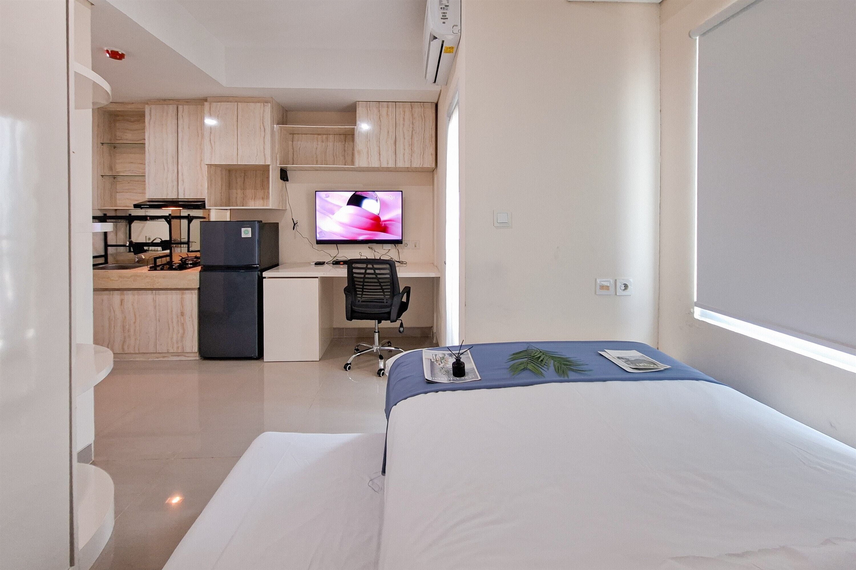 A compact hotel room with a bed, a desk with a computer and TV, a kitchenette, and a small office chair.
