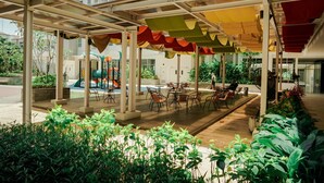 Un espace extérieur aménagé en terrasse, avec des tables et des chaises, entouré de verdure et avec une aire de jeux en arrière-plan.