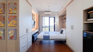 A narrow hotel room with a bed, a desk, and a balcony. - Cozy Studio Apartment At The Ayoma (South Tangerang)