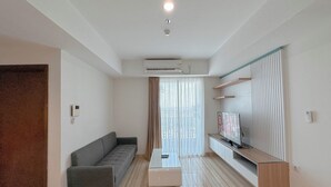 A modern living room with a grey sofa, a glass coffee table, a flat-screen TV on a wooden stand, and a large window with sheer curtains. - 3Br Near Mall At Grand Sungkono Lagoon Apartment (Surabaya)