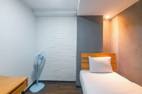 Une chambre d’hôtel avec un lit, une table de chevet en bois, un mur en briques blanches et un ventilateur sur pied.