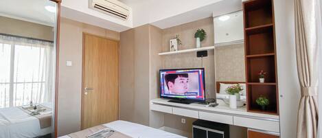 Habitación de hotel con cama, televisor, escritorio con computadora, estante con objetos decorativos y ventana con cortinas.