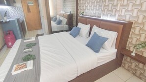 A neatly made bed with white and blue bedding, a bedside table with a magazine, and a mirror reflecting the room. - Studio Minimalist And Warm At Pavilion Permata Apartment (Surabaya)