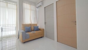 Una habitación moderna con un sofá color crema, una puerta de madera cerrada y una ventana con cortinas traslúcidas.