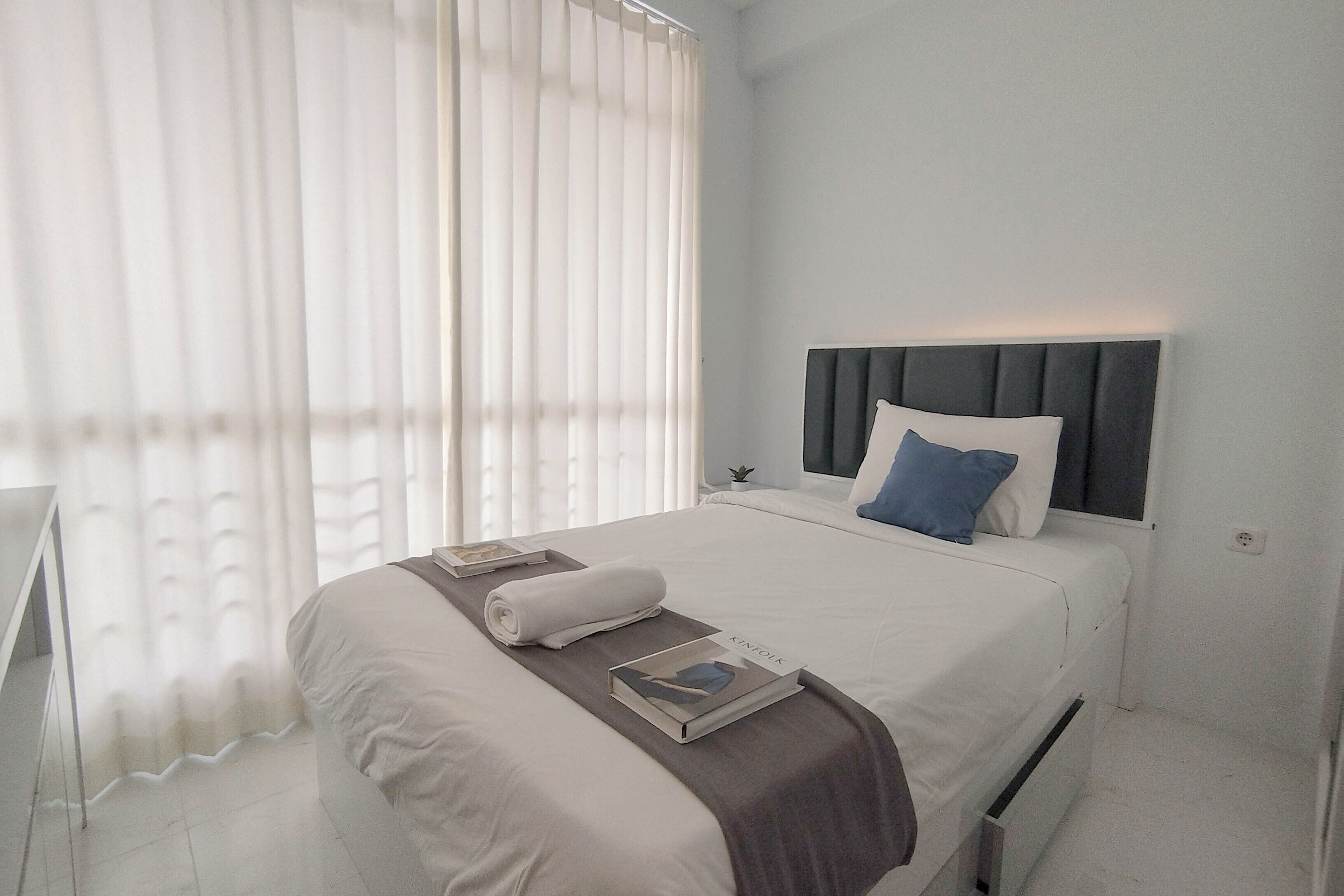 A modern hotel room with a neatly made bed, a bedside table, and a window with sheer curtains.
