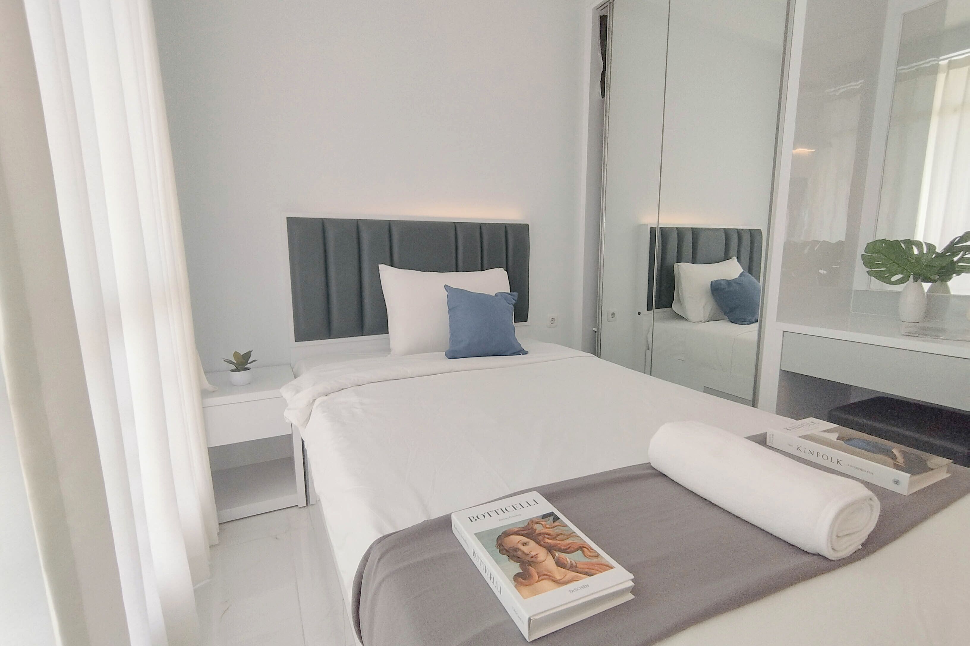 A modern hotel room with a neatly made bed, a small bedside table with a plant, and a large mirror reflecting the room.