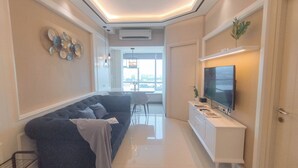 A modern living room with a dark-colored sofa, a white entertainment unit with a flat-screen TV, and a large window offering a city view. - Comfort 2Br Direct Access To Mall At Benson Supermall Mansion Apartment (Surabaya)