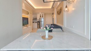 A modern kitchen with white cabinets, a stainless steel refrigerator, and a dining area with a marble table and a vase of flowers. - Comfort 2Br Direct Access To Mall At Benson Supermall Mansion Apartment (Surabaya)
