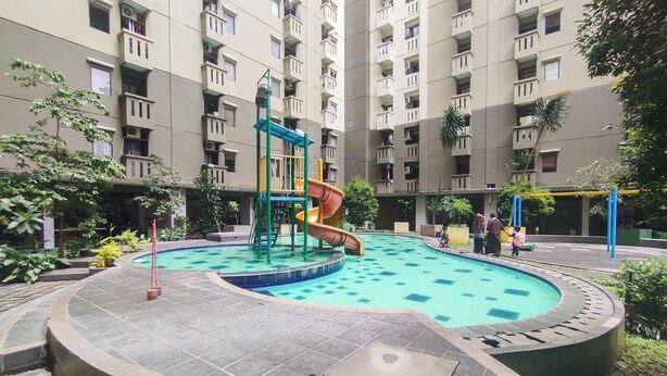 Una piscina con tobogán y estructura de juegos, rodeada por un área ajardinada con árboles y un edificio de varios pisos al fondo.