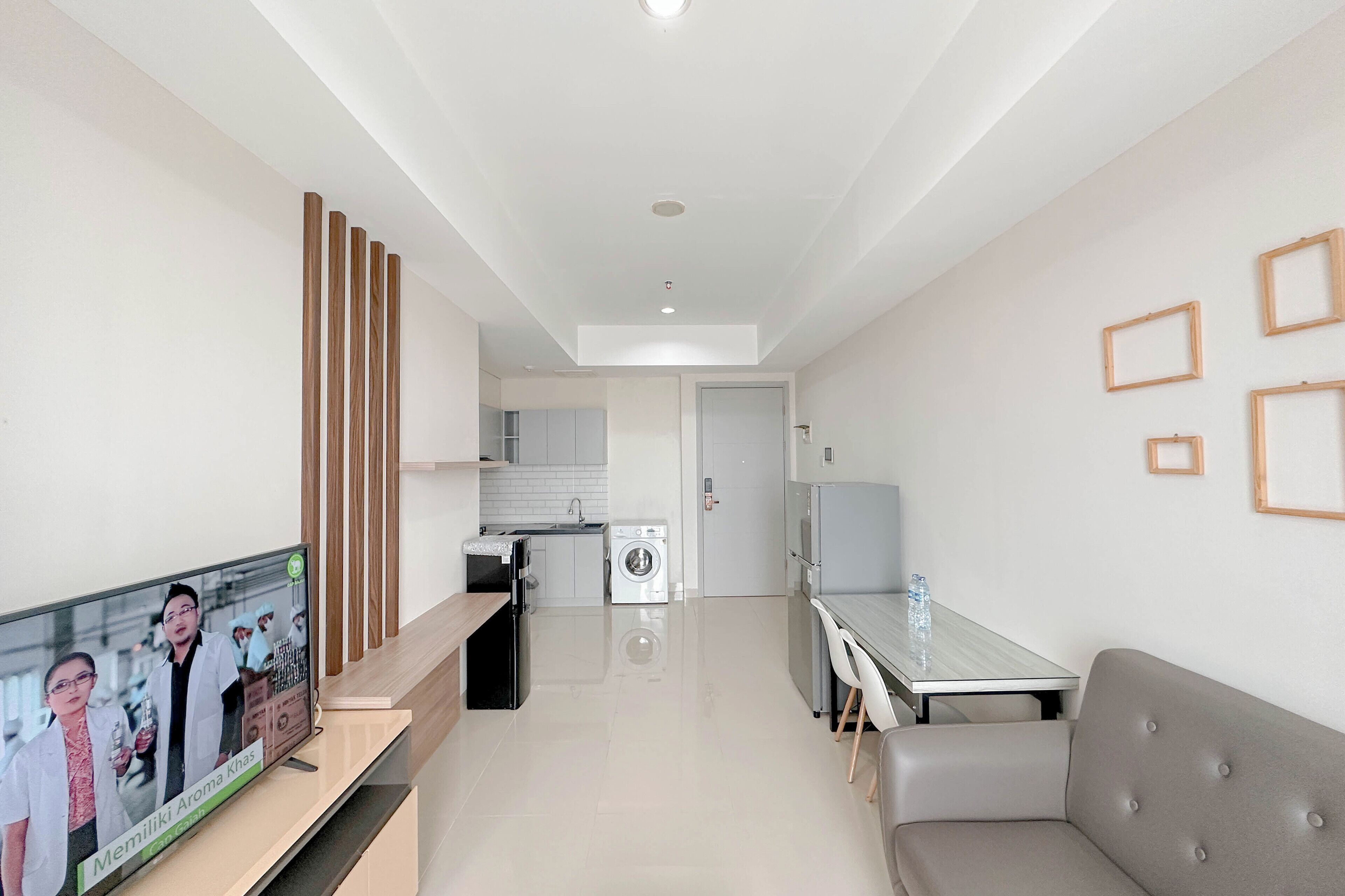 A modern living room with a flat-screen TV, a grey sofa, a dining table, and a kitchen in the background.