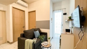 A modern living room with a sofa, a small round table, and a TV mounted on the wall. There are wooden doors and a window with curtains. - Minimalistic 2Br Near Tokyo Hub Apartment At Tokyo Riverside Pik 2 (Teluknaga)