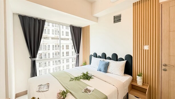 A modern bedroom with a large bed, a window with curtains, a wooden wardrobe, and a small plant on the bedside table. - Minimalistic 2Br Near Tokyo Hub Apartment At Tokyo Riverside Pik 2 (Teluknaga)