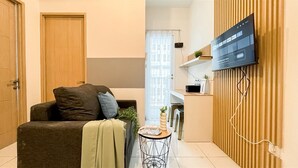 A modern living room with a sofa, a small side table, a television mounted on a wooden panel, and a door. - Minimalistic 2Br Near Tokyo Hub Apartment At Tokyo Riverside Pik 2 (Teluknaga)