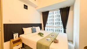 A bedroom with a bed, a nightstand, a window with curtains, and a view of buildings outside. - Modern 2Br Apartment Near Tokyo Hub At Tokyo Riverside Pik 2 (Teluknaga)