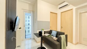 A modern living room with a sofa, a small table, and a TV mounted on the wall. There is a door leading to a balcony, and an air conditioning unit on the ceiling. - Modern 2Br Apartment Near Tokyo Hub At Tokyo Riverside Pik 2 (Teluknaga)