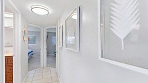 Ein Flur mit weißen Wänden, Fliesenboden und einer zur Schlafzimmertür. An der Wand befinden sich gerahmte Blattform-Motive und eine kleine Holzkommode links.