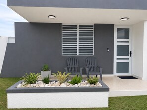 A modern building exterior with a grey facade, a white door with glass panels, two grey Adirondack chairs, and a planter with various plants. - Casa MTOS Relax & Explore near San Juan & Beaches (Carolina)