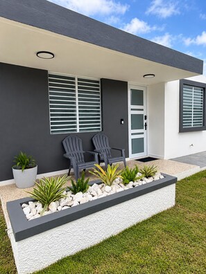 Une maison moderne avec un toit plat, un porche à l’avant doté de deux chaises et un jardinier contenant des plantes et des roches.