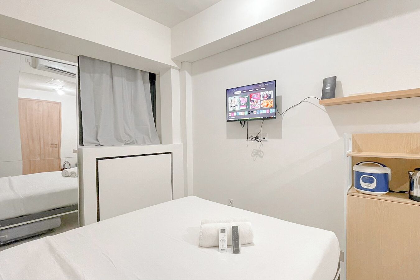 A hotel room with a bed, a TV mounted on the wall, a bedside table with a remote control, and a wooden shelf with a kettle.