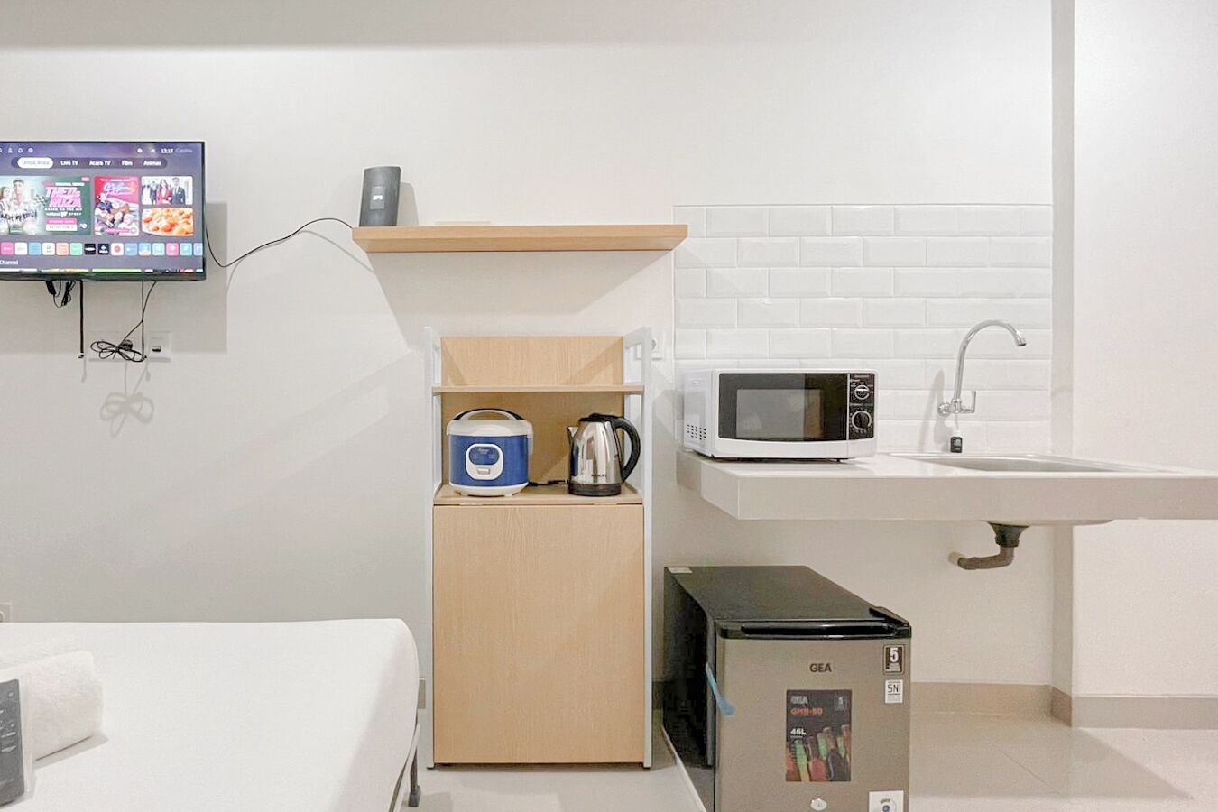 A compact kitchenette with a microwave, kettle, and a small refrigerator. A TV is mounted on the wall displaying various channels.