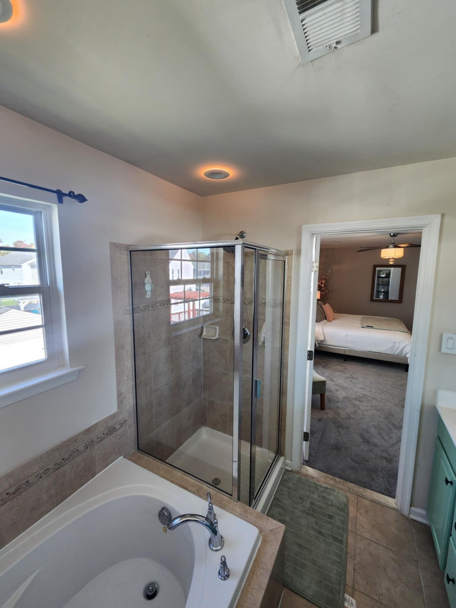 A modern bathroom with a glass-enclosed shower, a large bathtub, and a visible bedroom with a bed and a ceiling fan.