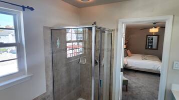A modern bathroom with a glass-enclosed shower, a large bathtub, and a visible bedroom with a bed and a ceiling fan.