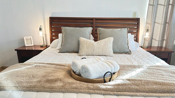 Una cama bien hecha con un edredón beige suave, dos almohadas y una bandeja de madera con toallas dobladas.