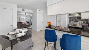 Una cocina moderna con armarios blancos, encimera oscura y campana extractora de acero inoxidable. Hay un comedor con mesa y sillas de vidrio, y una silla azul en primer plano.