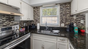 Una cocina moderna con armarios blancos, estufa de acero inoxidable, encimera negra, ventana con persianas y un salpicadero estampado.